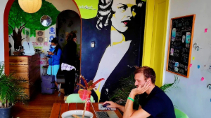 Hostel guest sitting at the big table in the living room; hostel guests doing check-in at the reception desk behind; bright colors in the living room and reception.